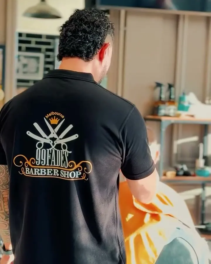 A 99 Fades barber at work, the shop crest printed on the back of his shirt